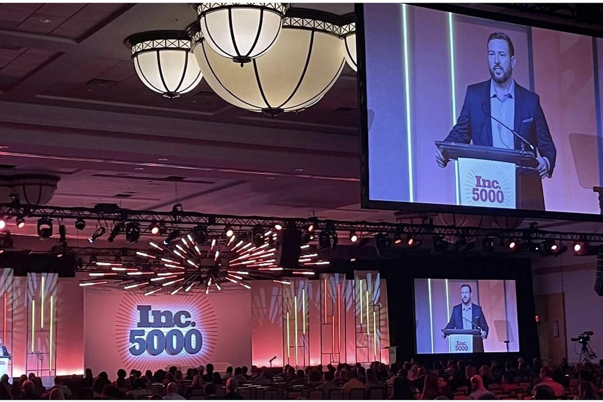 RJ Blake speaking from a podium on the main stage of the Inc. 5000 conference, shown on large screens above a full audience.