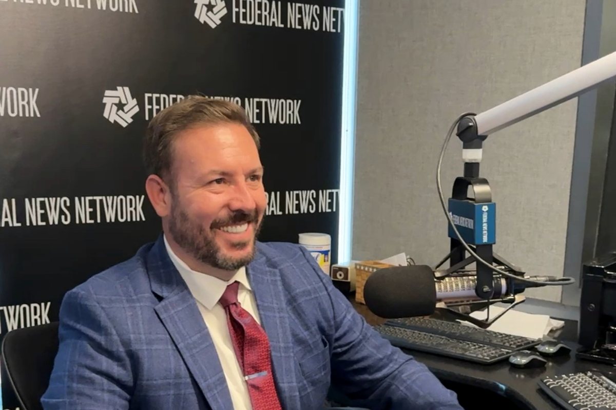RJ Blake smiling at a broadcast desk in front of a Federal News Network backdrop, with a studio microphone beside him.