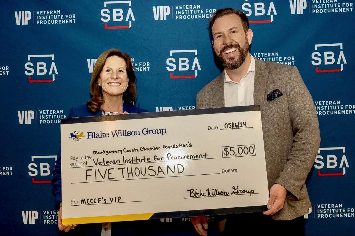 RJ Blake holding an oversized $5,000 check from Blake Willson Group presented to the Veteran Institute for Procurement, alongside a representative, in front of a VIP and SBA backdrop.
