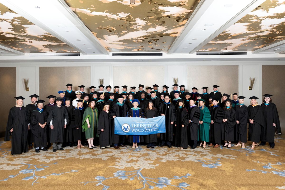 A commencement class photo of graduates and faculty in academic regalia behind an Institute of World Politics banner.