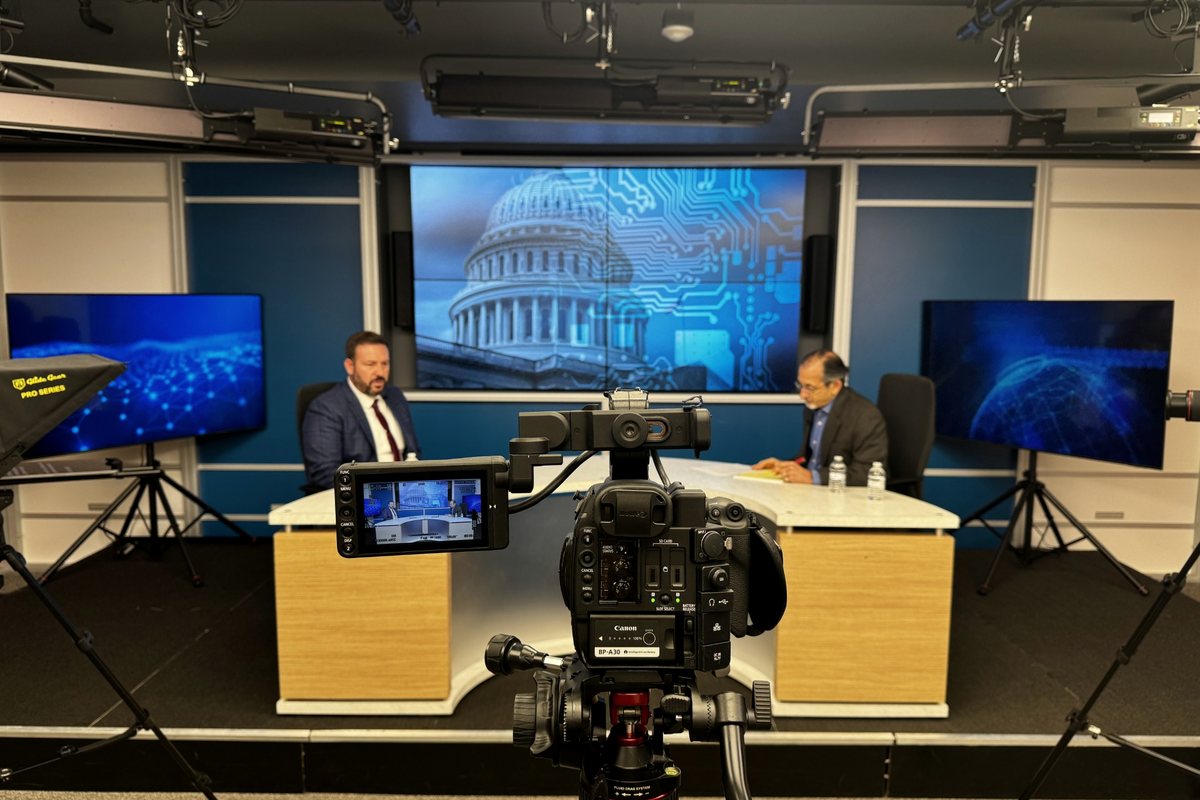 A television studio seen from behind the camera, with RJ Blake and an interviewer seated at the anchor desk in front of a Capitol dome graphic.