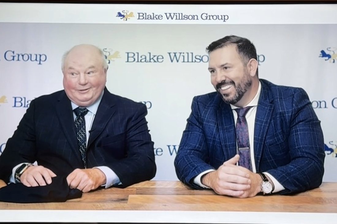 RJ Blake seated at a table with a guest in front of a Blake Willson Group branded backdrop during a recorded conversation.