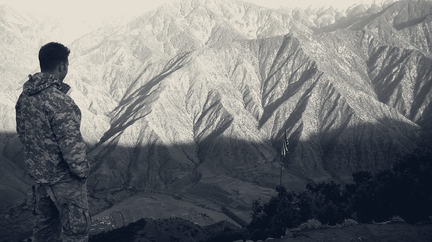 A soldier stands at an observation post looking across a wide Afghan valley toward terraced fields and steep mountain ridgelines, with a U.S. flag flying below.