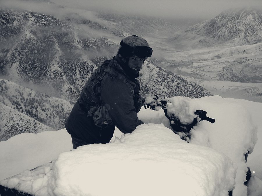 A soldier in goggles clearing heavy snow from equipment on an exposed ridgeline above a snow-covered valley.