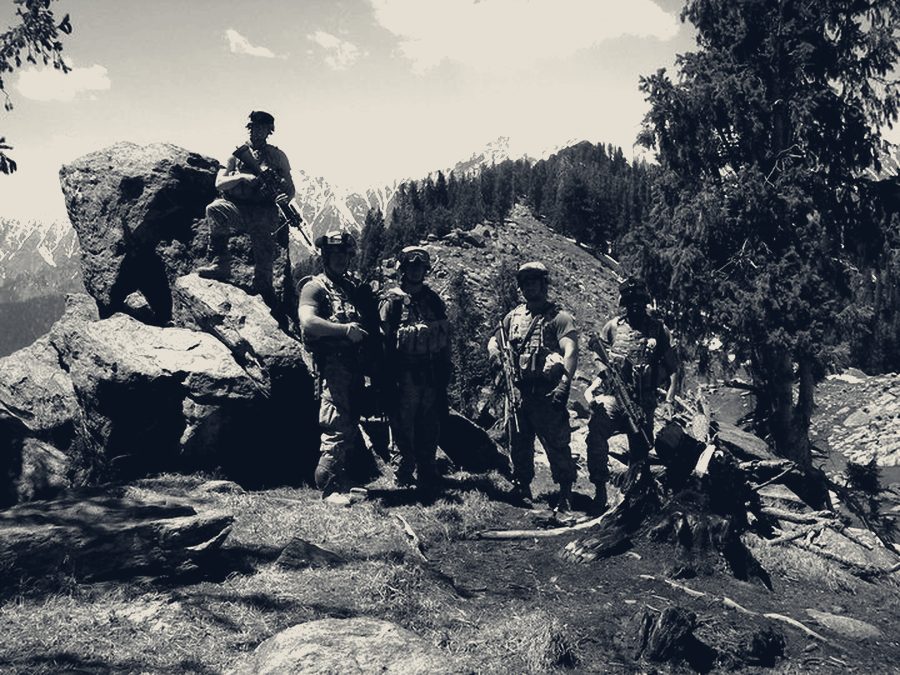 A reconnaissance element halted among boulders and pines with snow-capped peaks behind them.