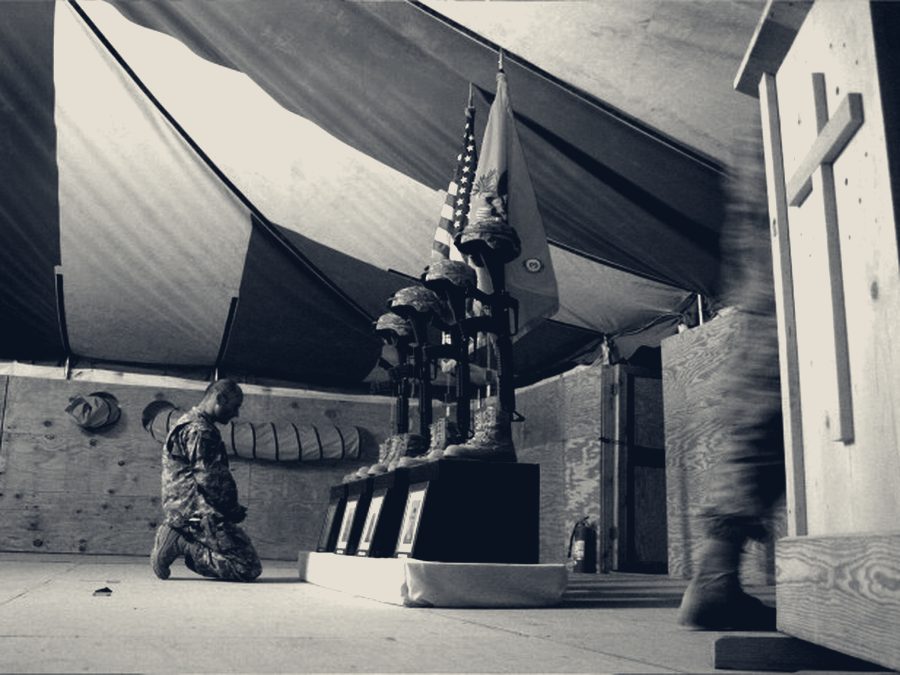 A soldier kneels before a memorial of inverted rifles, helmets and boots arranged beneath the U.S. and unit colors at a forward operating base.